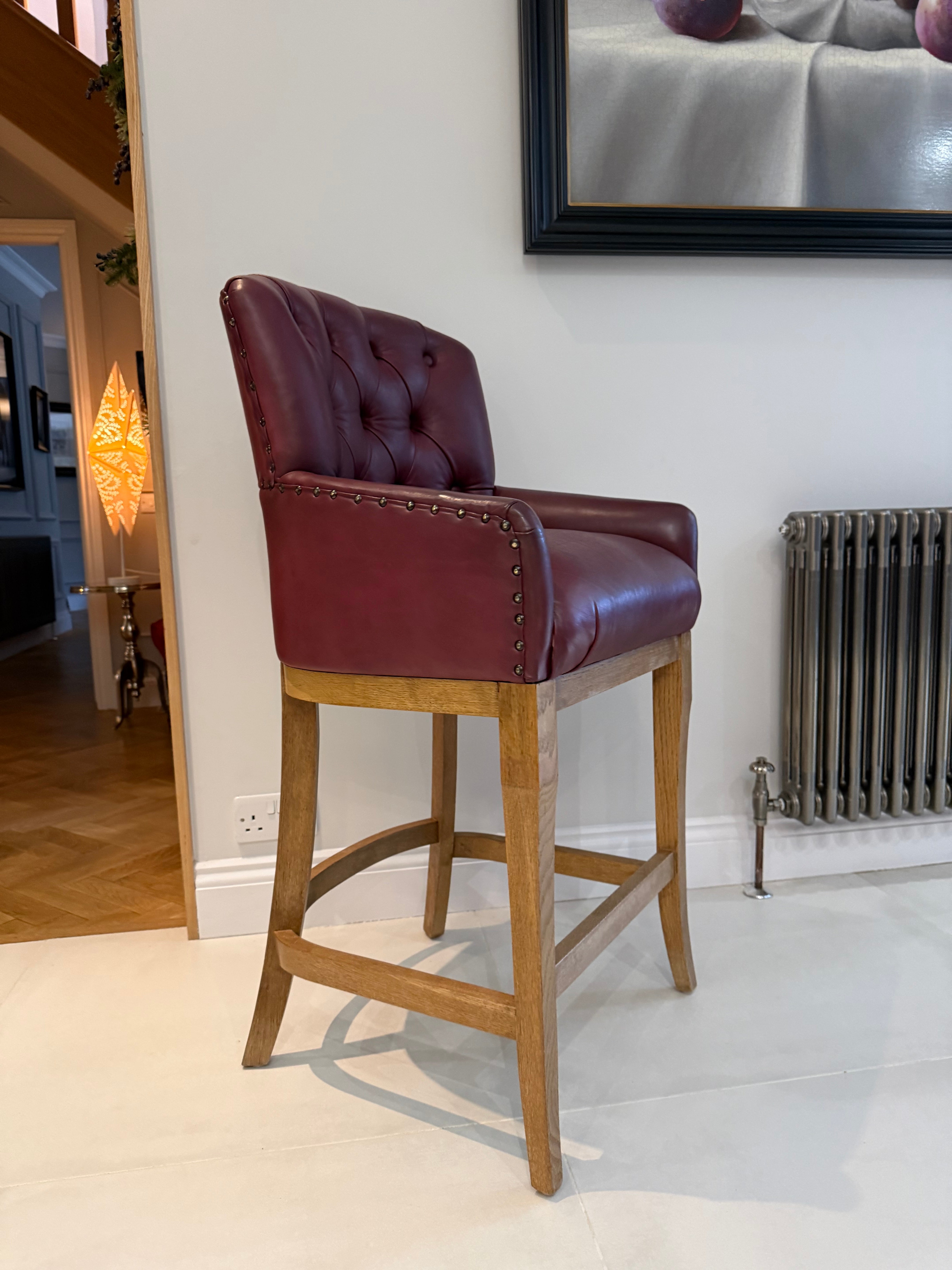 A single-tufted bar stool with burgundy leather upholstery and a natural wood frame, positioned in a modern home.