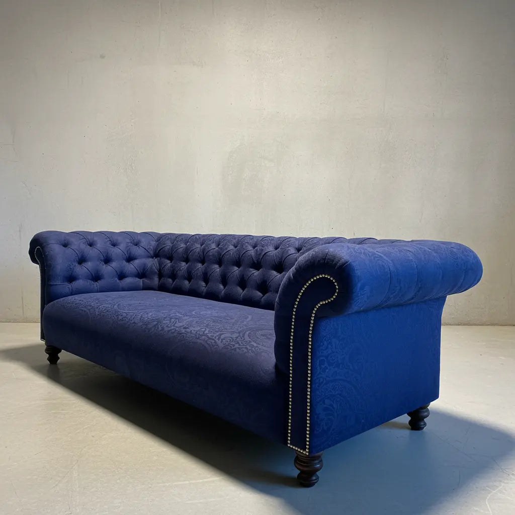 A classic Chesterfield-style daybed in deep blue velvet with tufted backrest, rolled arms, nailhead trim, and dark wooden legs, photographed in a minimalist studio setting.