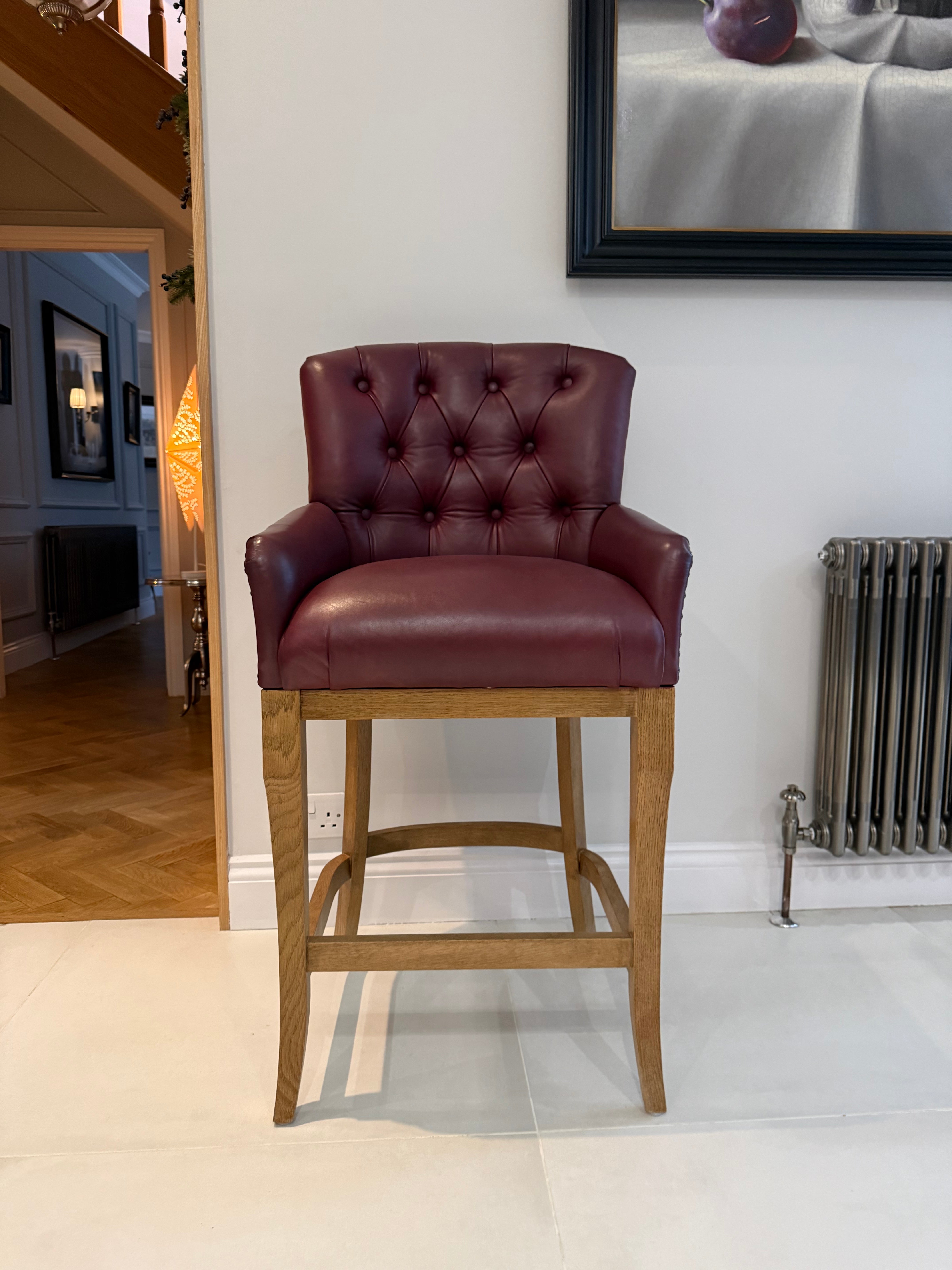 A tufted leather bar stool with a deep burgundy seat and backrest, mounted on a light oak wooden base, set in a home interior.