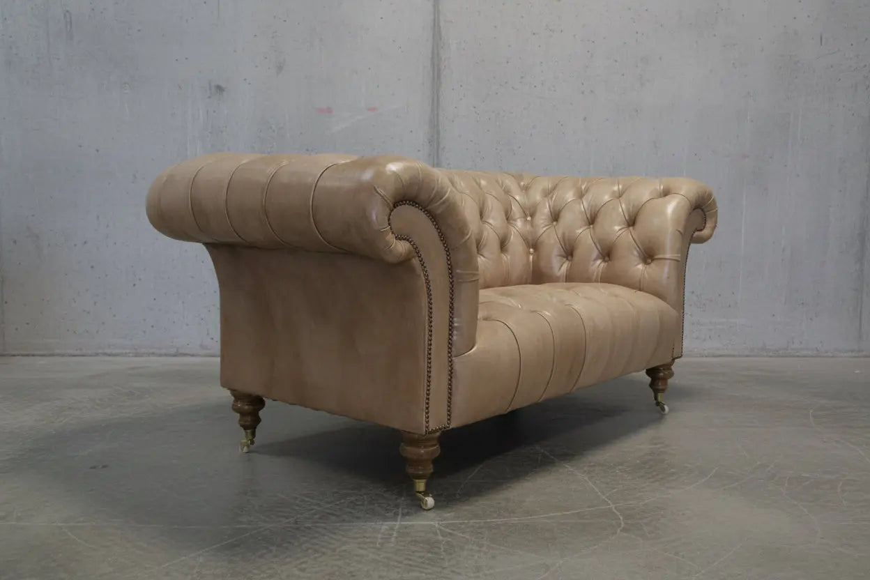 A classic Chesterfield loveseat sofa upholstered in tan leather, featuring deep button tufting, rolled arms, and turned wooden legs with brass casters, set in a bright room with white floorboards and a light gray wall.