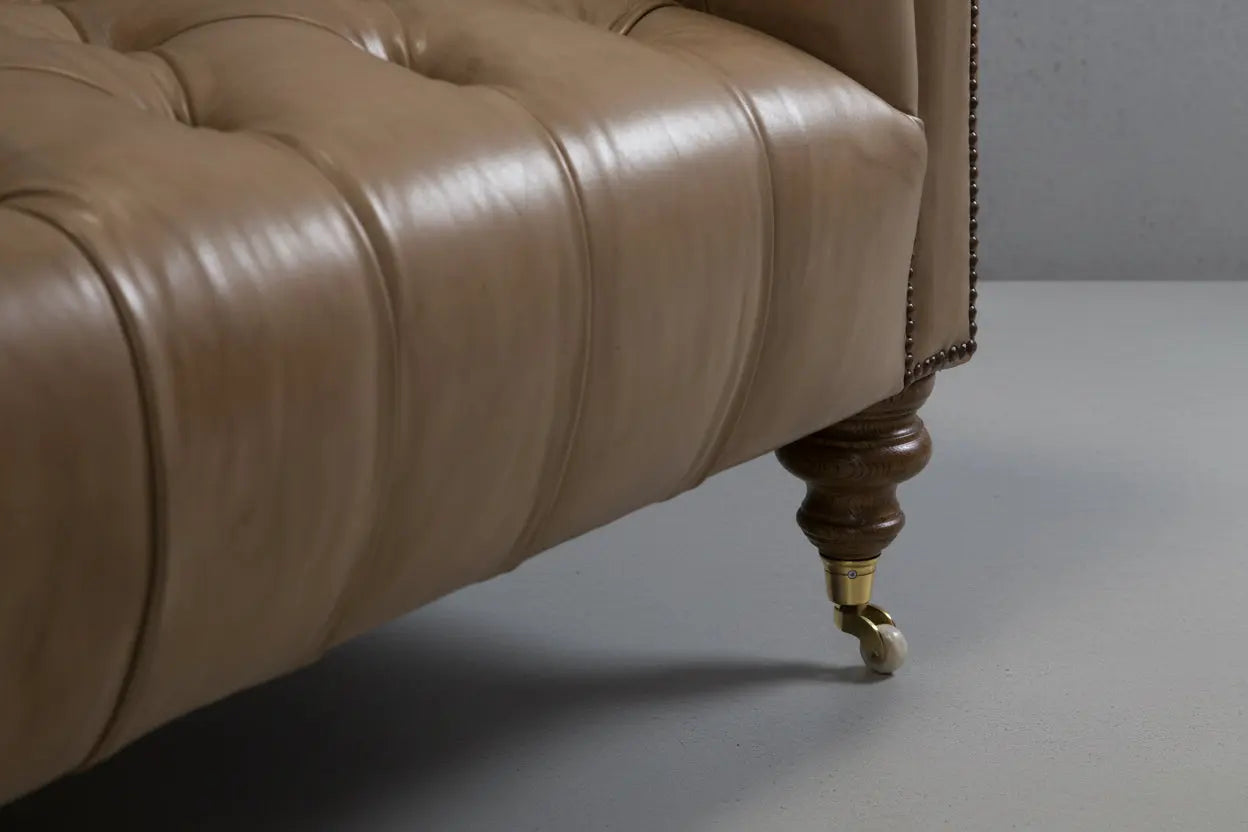 Close-up of a tufted leather chaise lounge with a dark wood turned leg and brass caster wheels, set on a white-painted wooden floor.