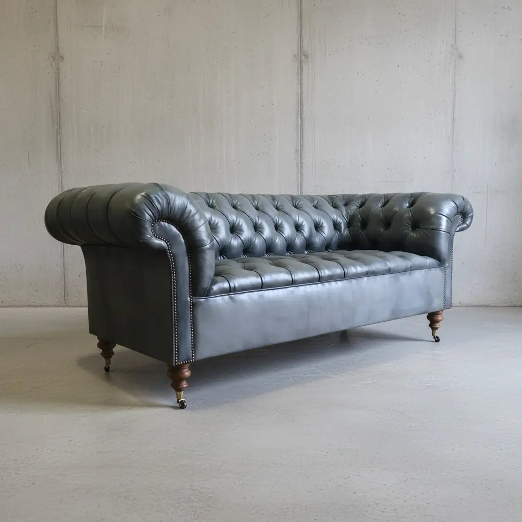 A classic Chesterfield loveseat sofa upholstered in dark charcoal gray leather, featuring deep button tufting, rolled arms, nailhead trim, and turned wooden legs, set against a concrete wall and floor.