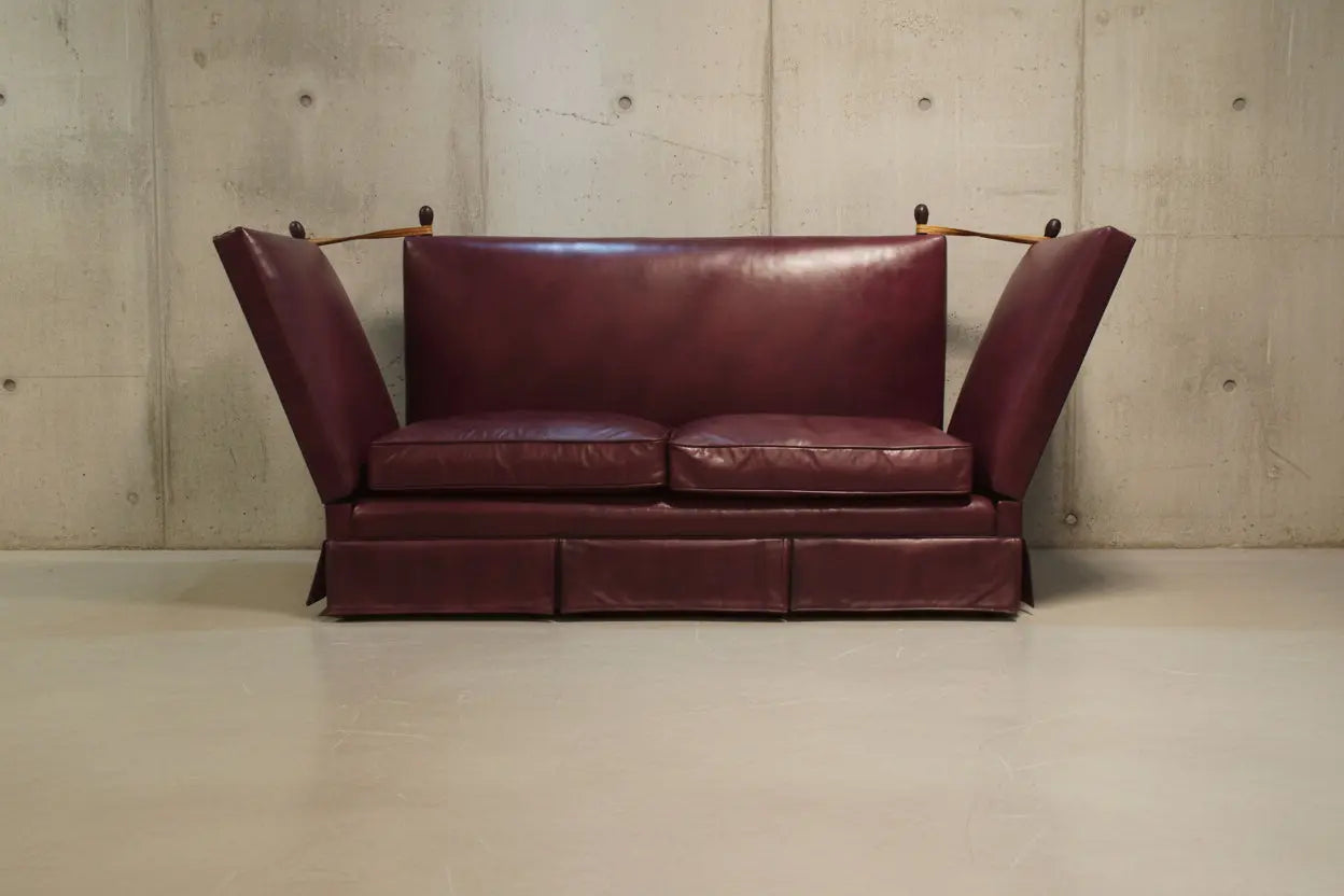 A mid-century modern  style knoll leather loveseat sofa in a deep burgundy or maroon color, featuring angled armrests with small wooden supports and a low boxy base, set against a neutral beige wall and white wooden floor.